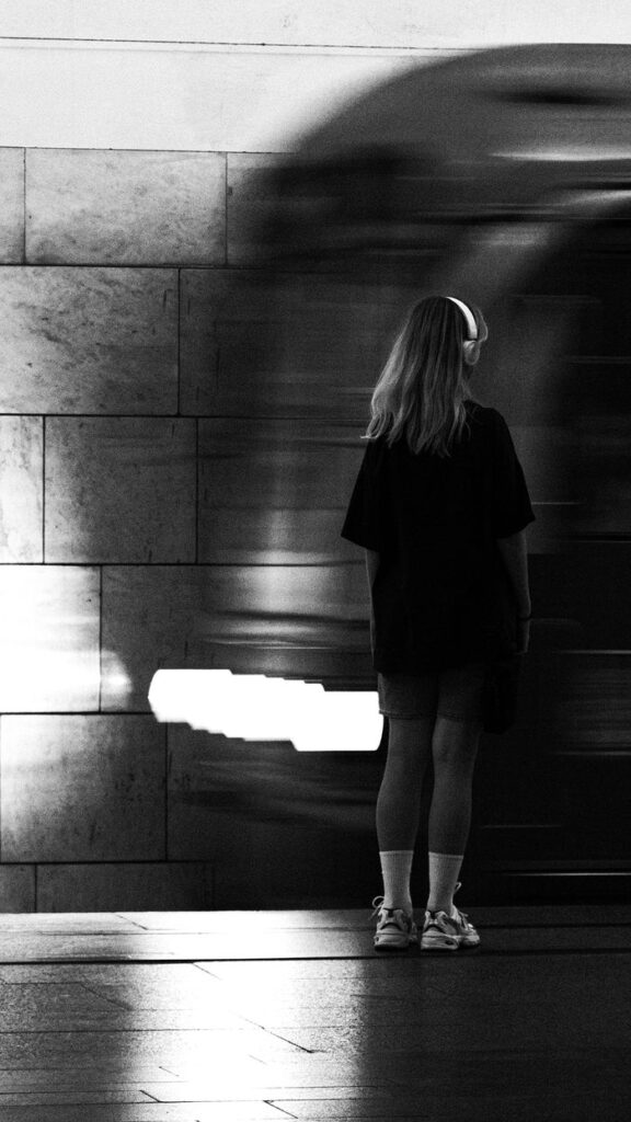 A woman in headphones stands at an urban subway platform as a train speeds by in black and white.
