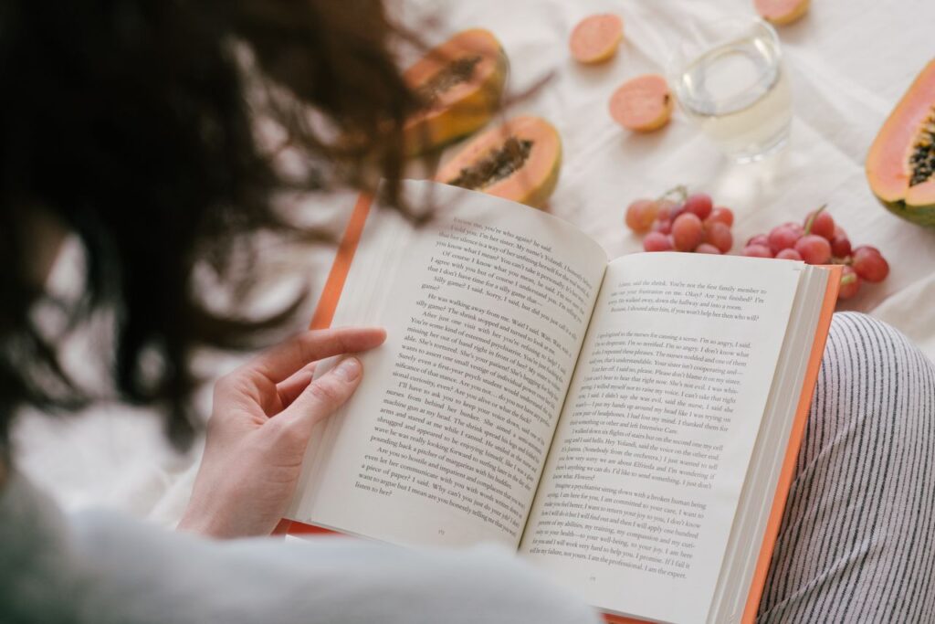 A person enjoys reading a book surrounded by fresh papaya, grapes, and a drink, creating a cozy indoor scene.