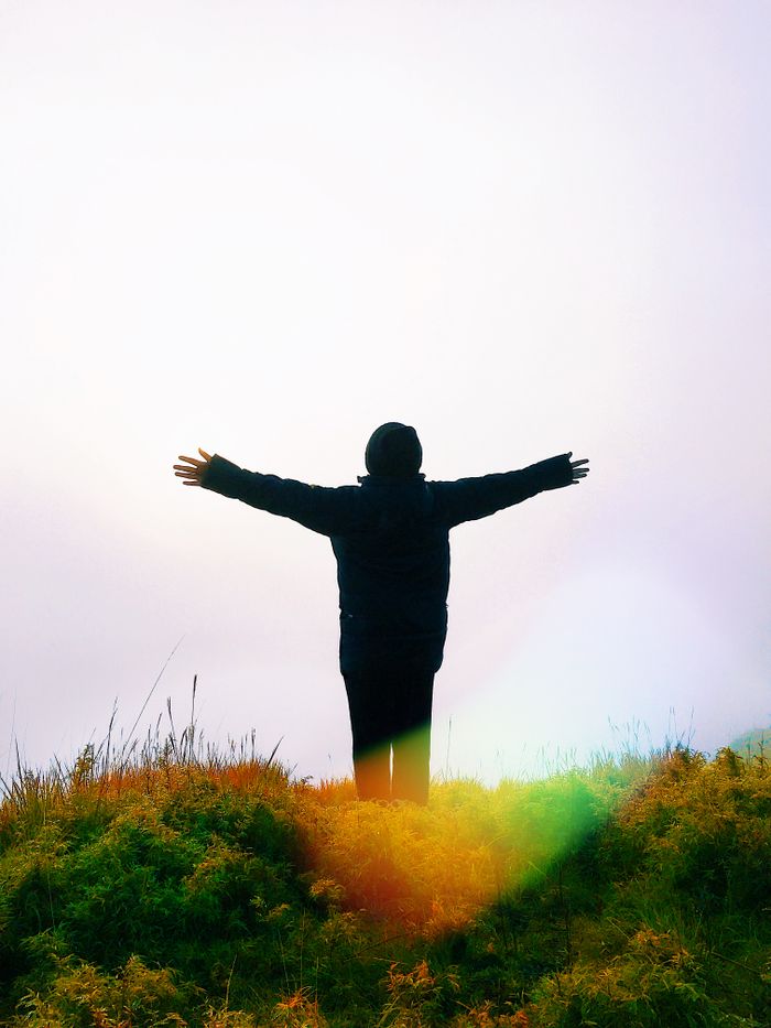 A silhouetted man stands with arms outstretched on a grassy hill, capturing a moment of freedom and connection with nature.